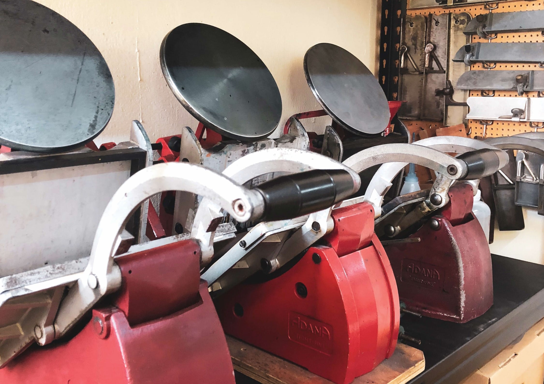Three red tabletop printing presses are lined up in a row with hand tools hanging on a pegboard in the background.