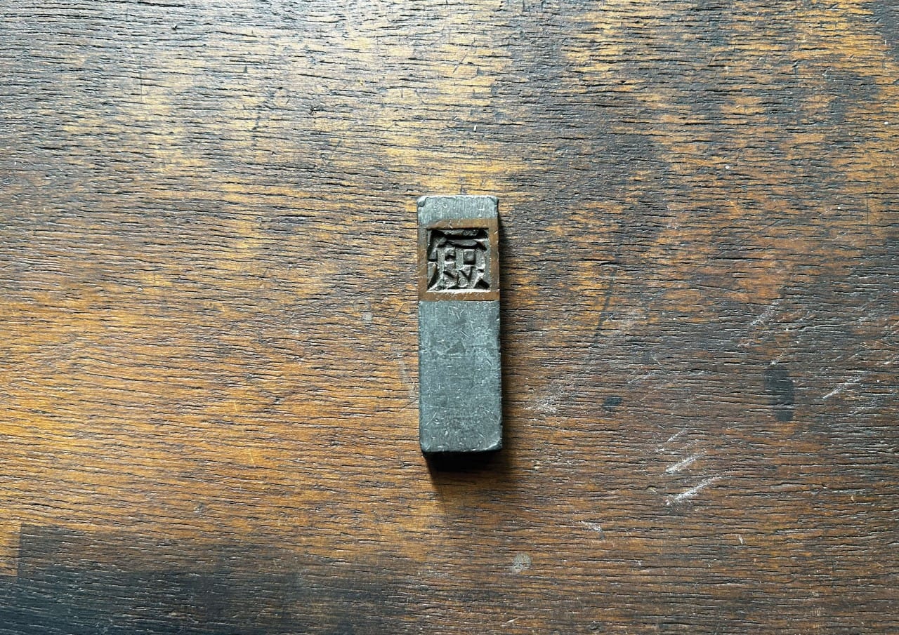 A single metal printing block with an engraved symbol is placed on a worn wooden surface.