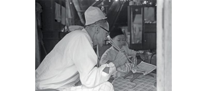 An elderly man wearing a cap and glasses sits inside a room, reading a newspaper with a pipe in his hand.