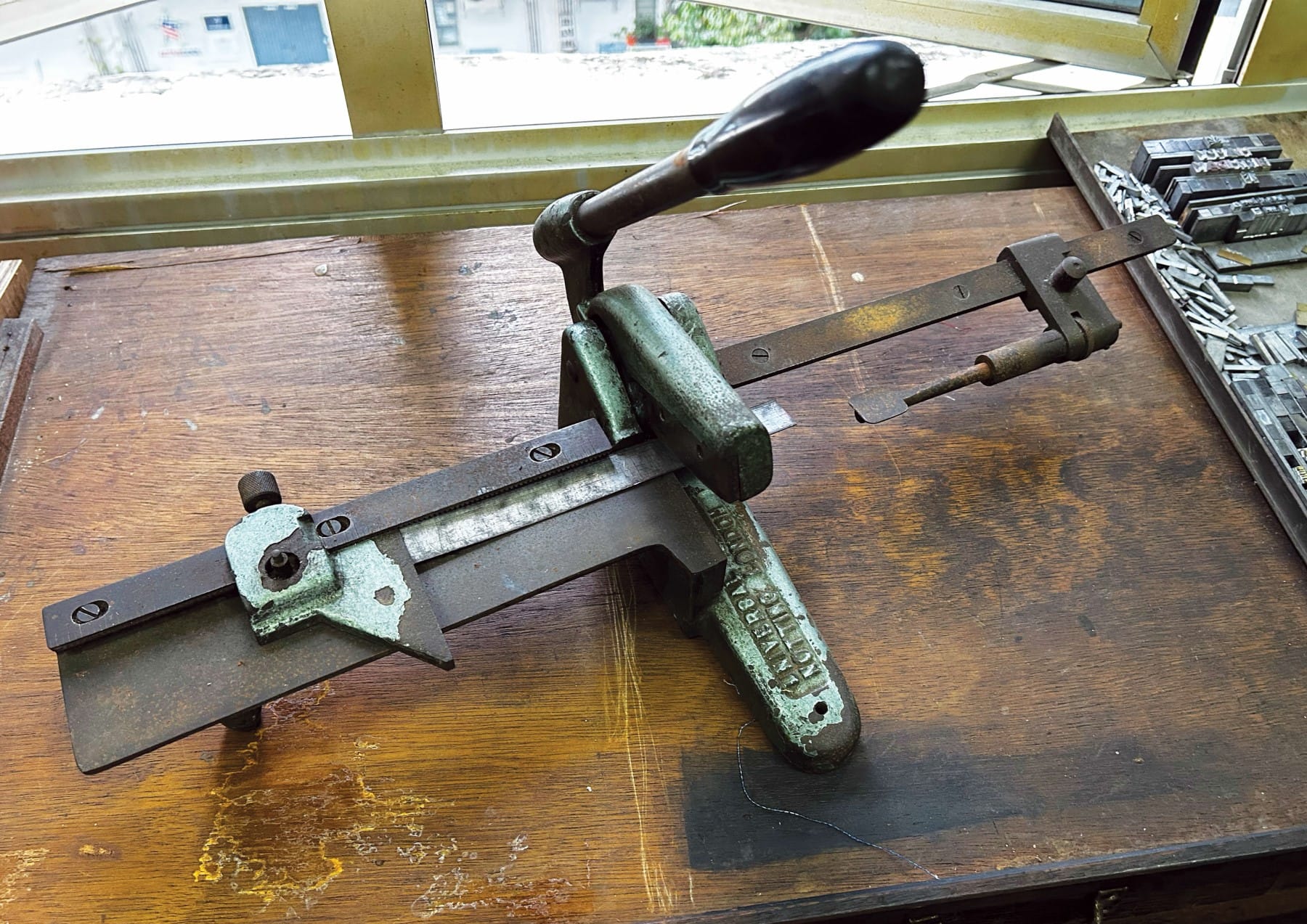 An old manual printer's tools on a wooden table near a window, featuring a type holder and metal type pieces.