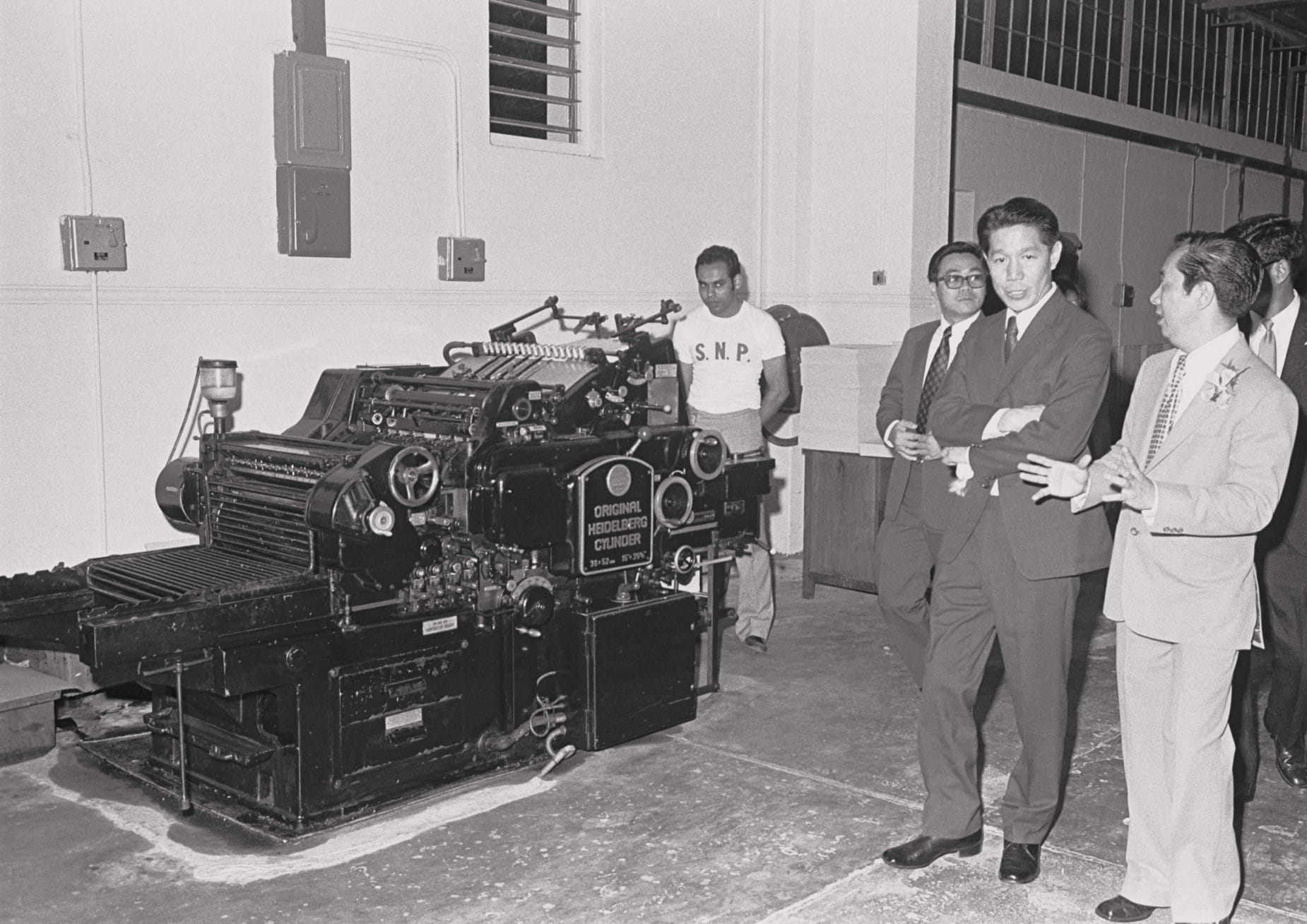Several men stand near a large industrial machine labeled "Original Heidelberg Cylinder" in a workshop environment.