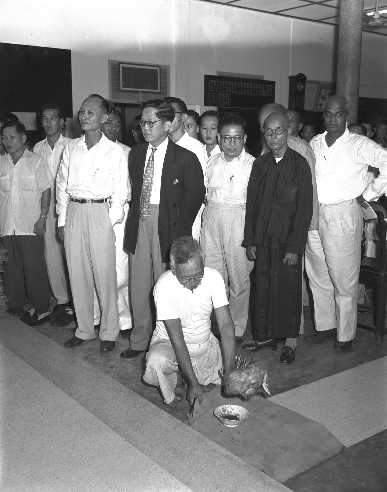 A man kneels down in front of a group of people, with a chicken and a bowl on the ground.