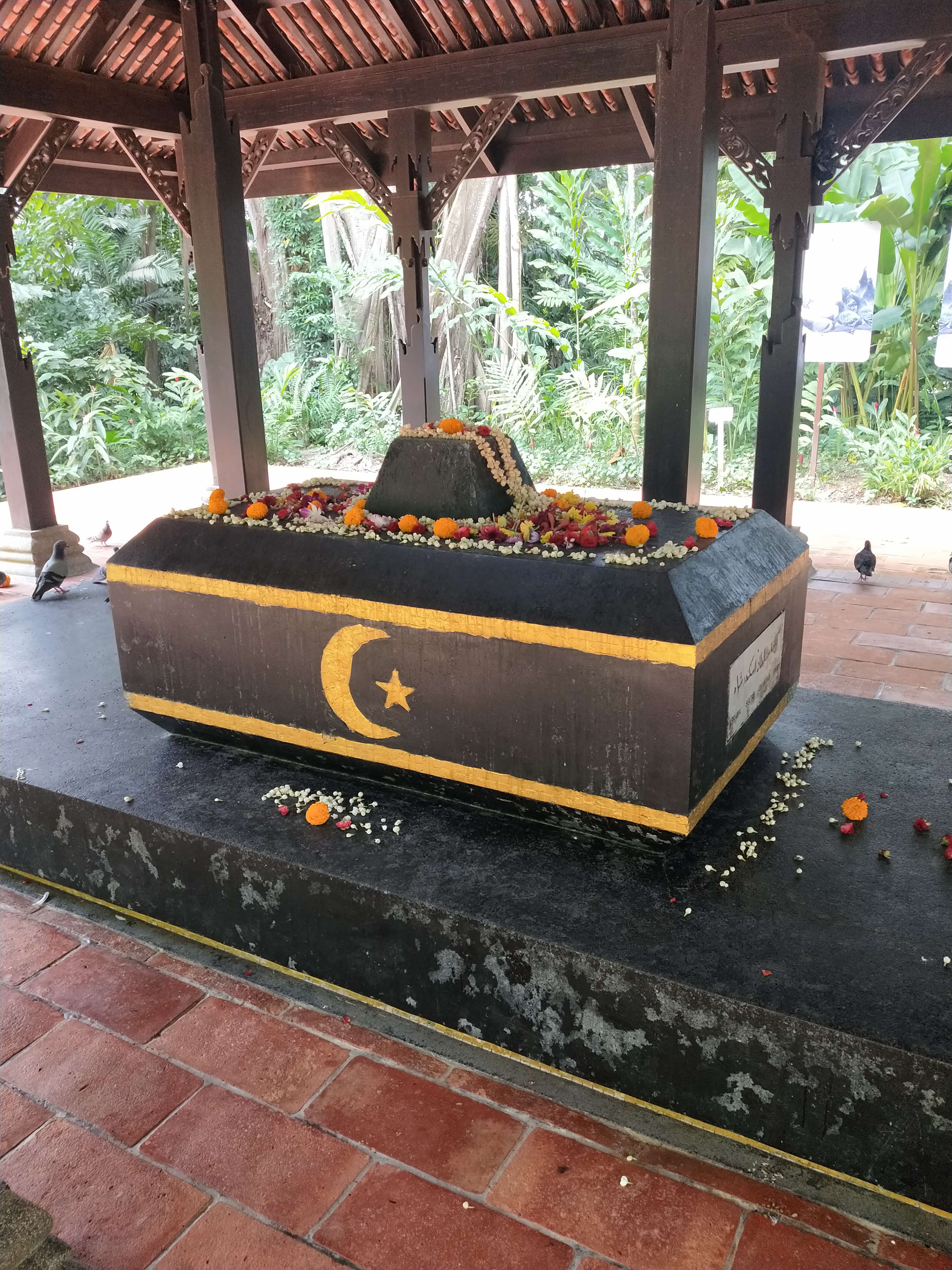 Outdoor tomb with yellow crescent and star, adorned with flowers, under a wooden pagoda surrounded by greenery.