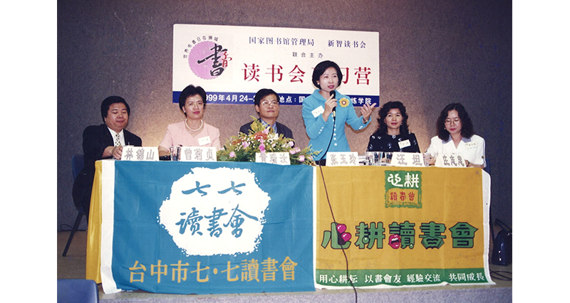 A group of people sit at a table with two banners featuring Chinese text and a flower arrangement.
