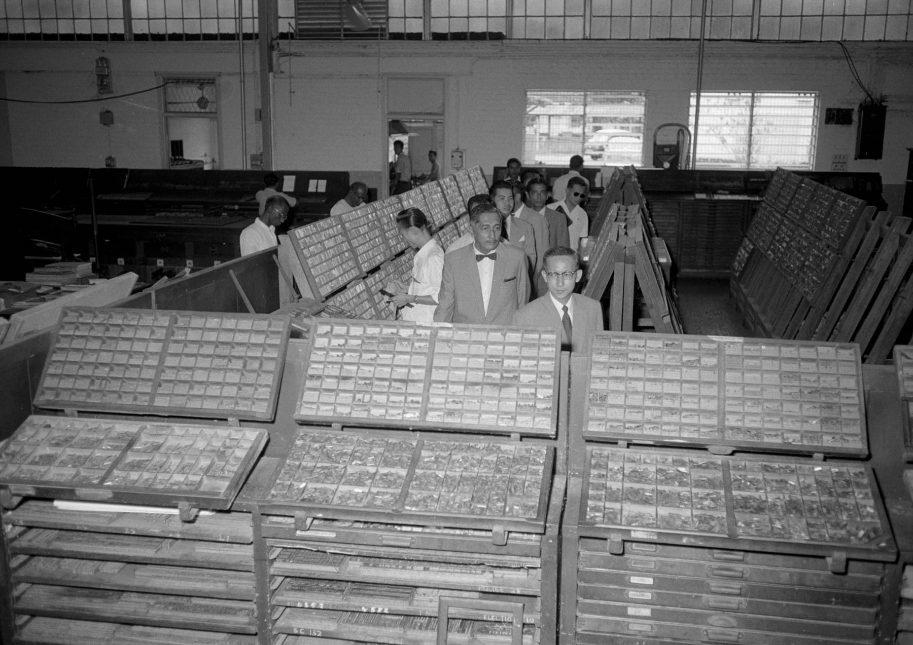 People standing between large racks of typefaces in an old printing workshop with windows in the background.