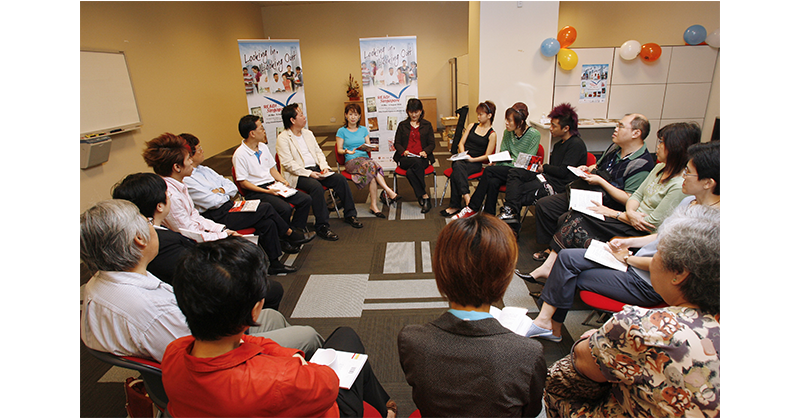 A group of people seated in a circle, holding papers, with two posters and colorful balloons on the background wall.