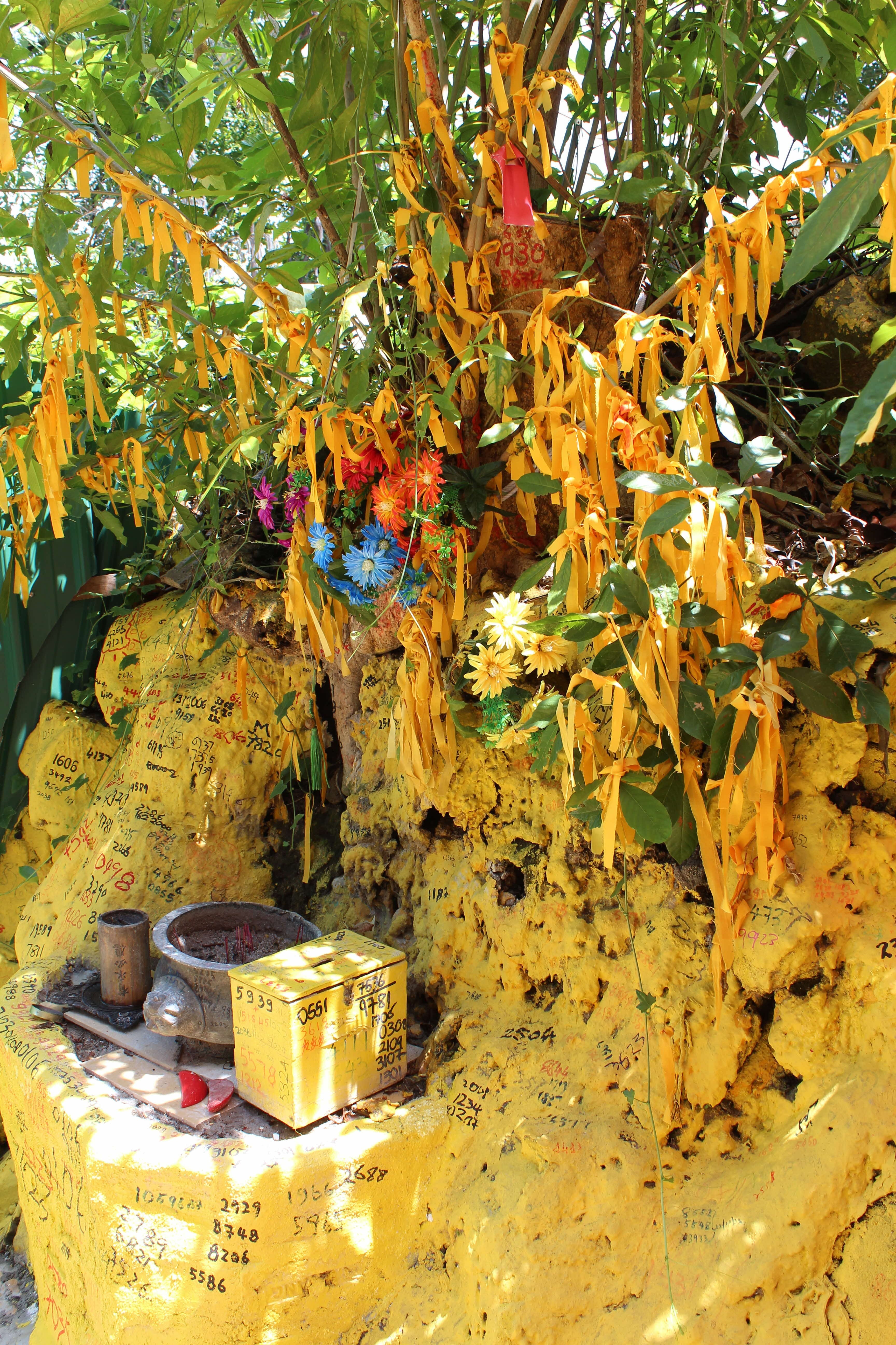 The tree near the keramat is adorned with ribbons, and lottery numbers are scrawled over the painted rocks, 2022. Courtesy of William L. Gibson.