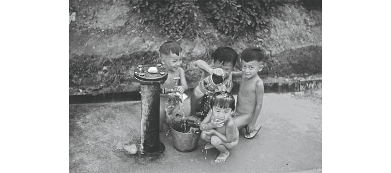 Several children are playing with water from a street pump, filling a bucket on a concrete surface.