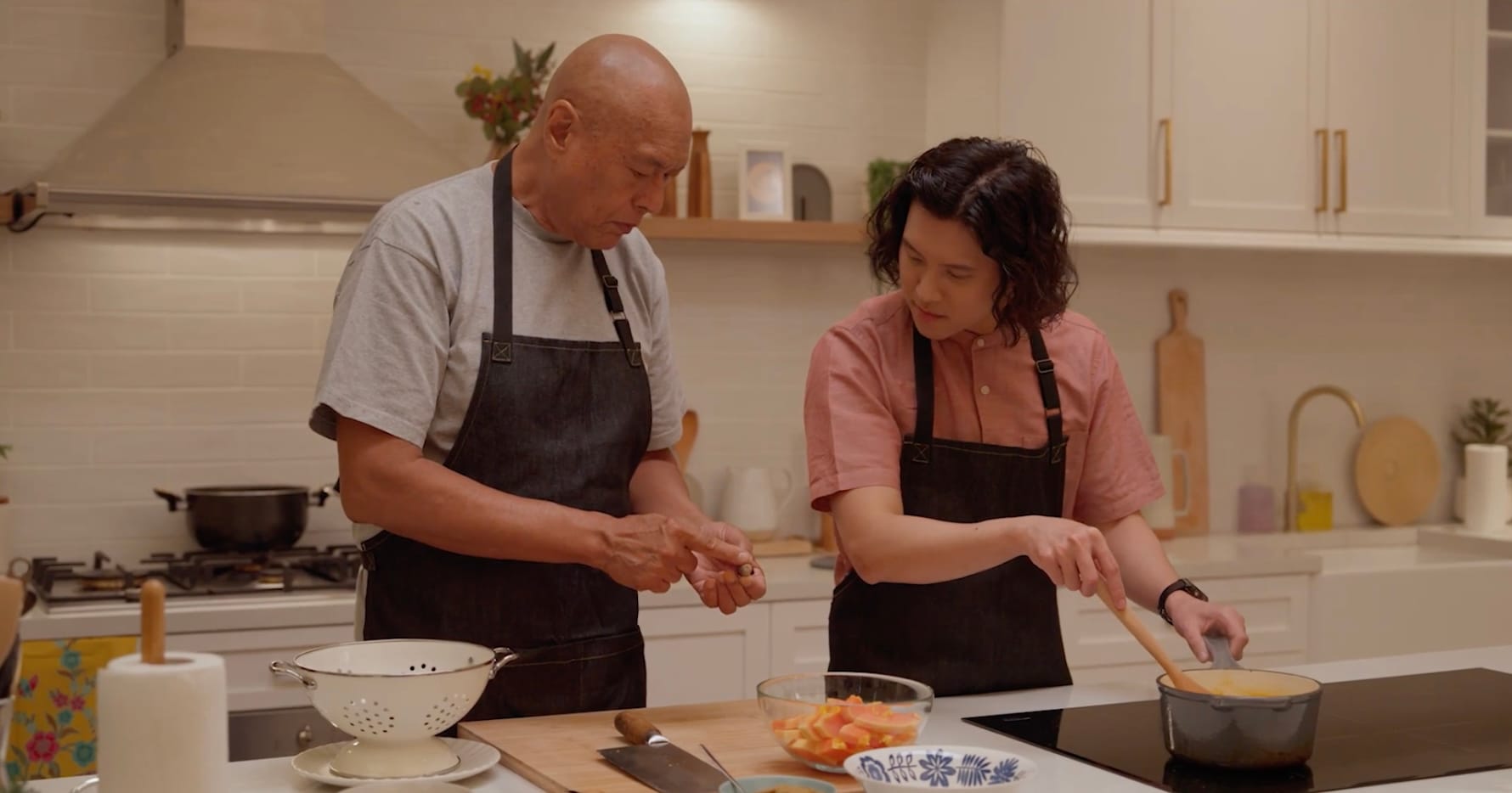 Two individuals in a kitchen are preparing food; one stirs a pot on the stove while the other handles vegetables.