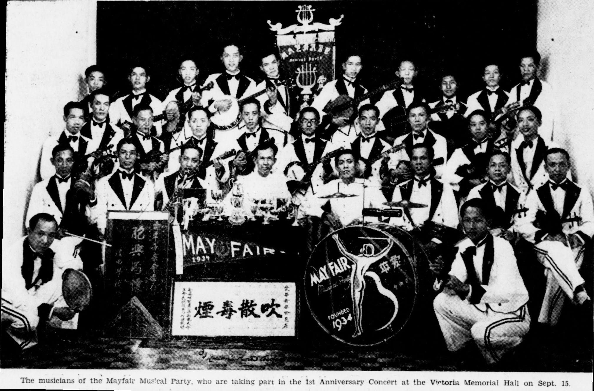 A large musical group in white and black outfits poses with instruments, behind banners reading "Mayfair" and Chinese characters.