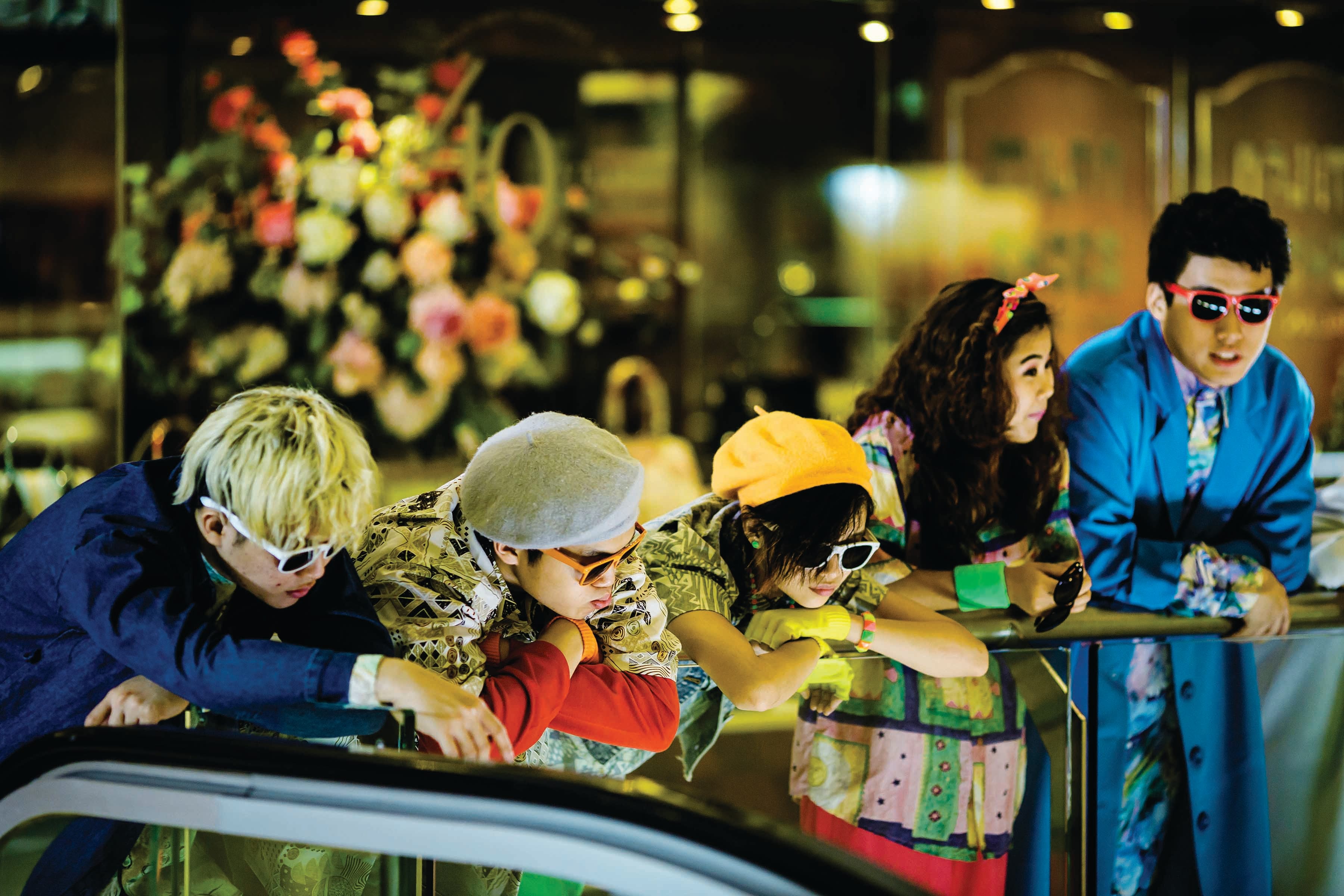 A group of people wearing colorful and eccentric clothing lean on a railing, with a floral arrangement in the background.