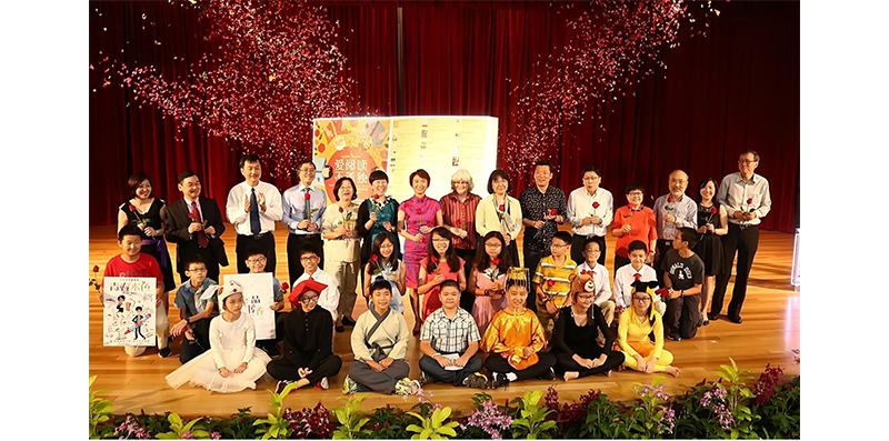 A large group of people in colorful outfits posing on stage with confetti and flowers.