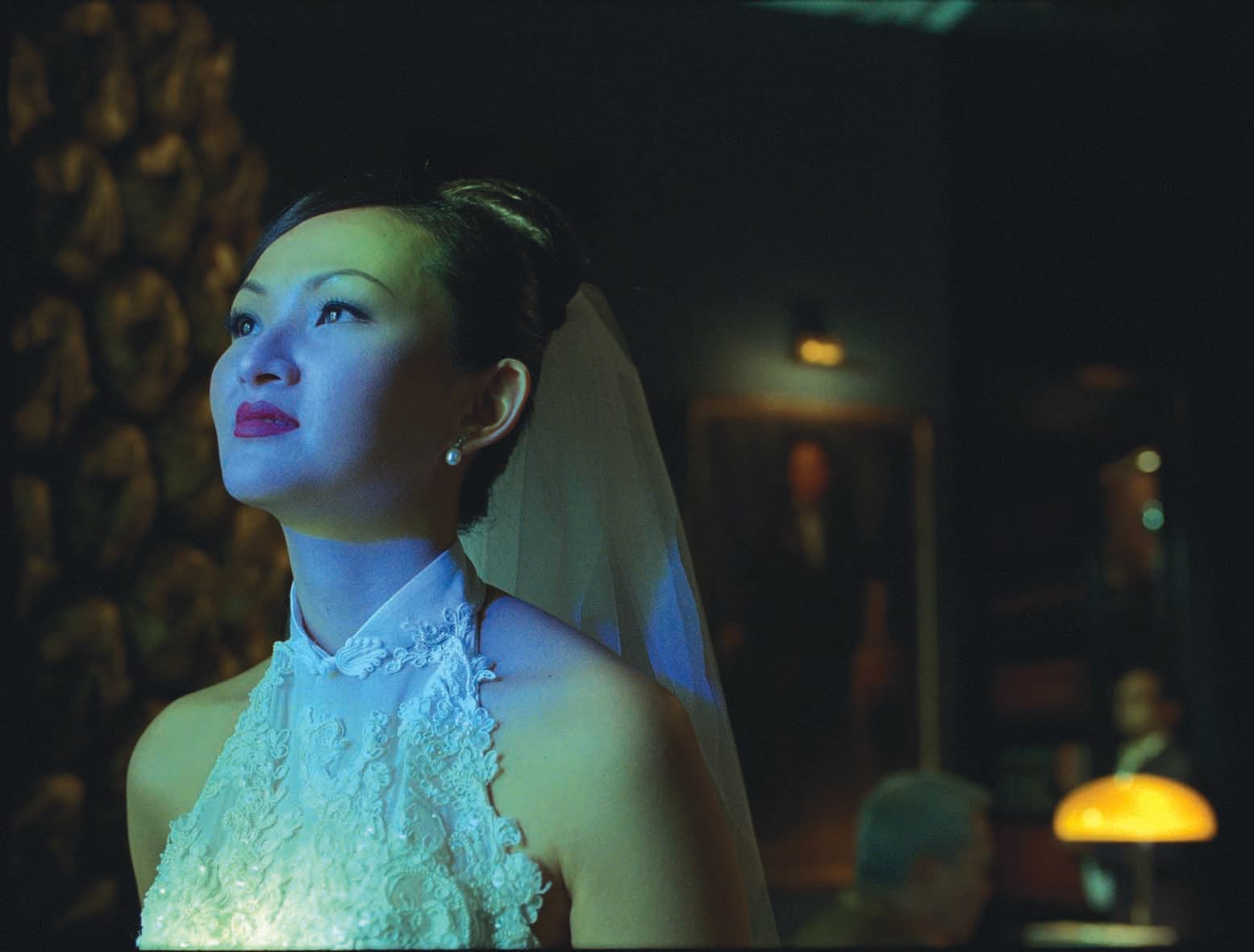 A person wearing a white embroidered dress and veil is illuminated by blue and green lighting in a dark room.