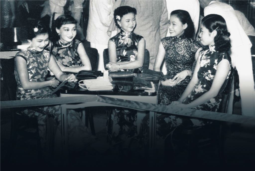 Five women wearing floral dresses are seated around a table with handbags.