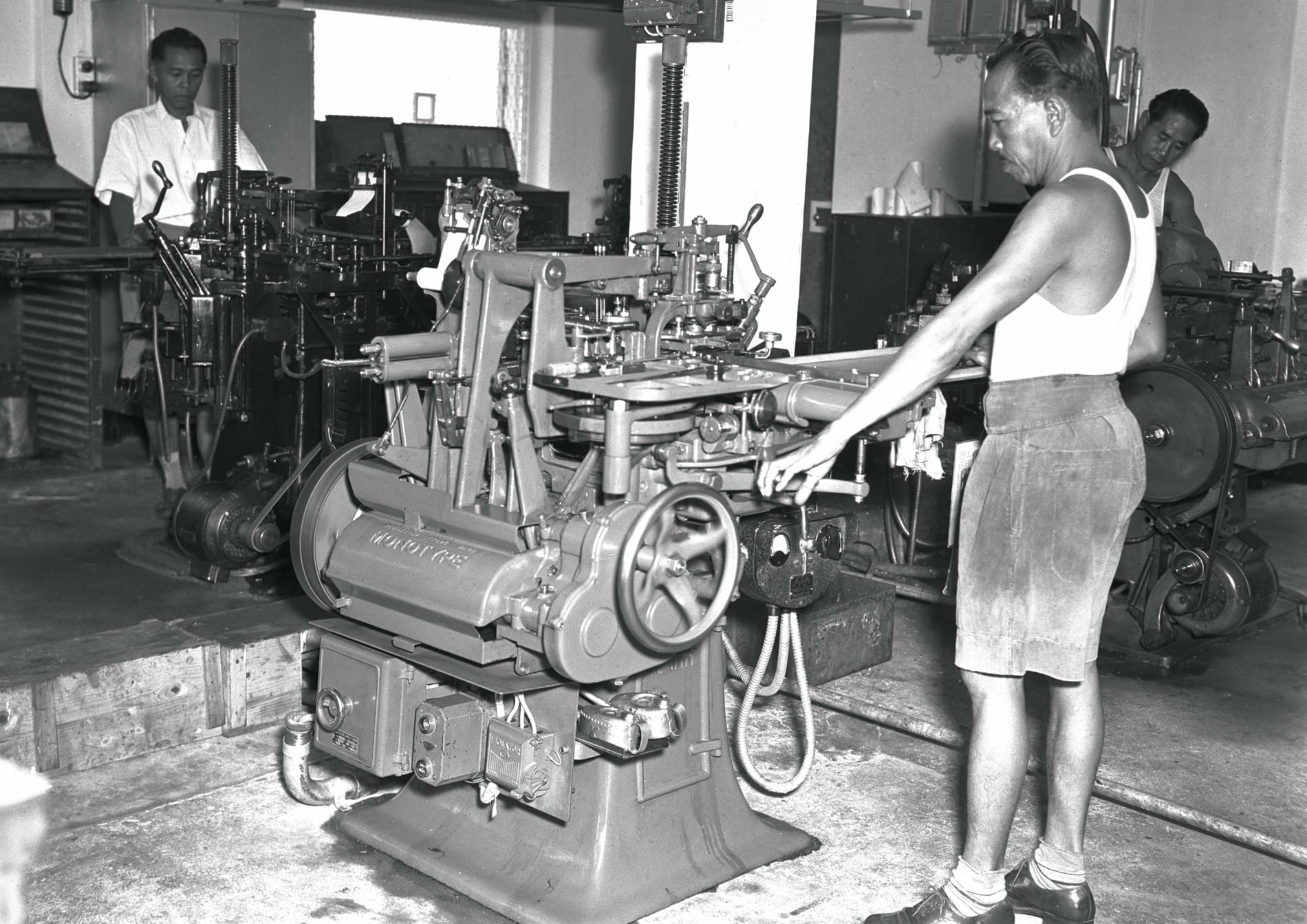 Several workers operate machinery in a printing workshop with mostly metal equipment.