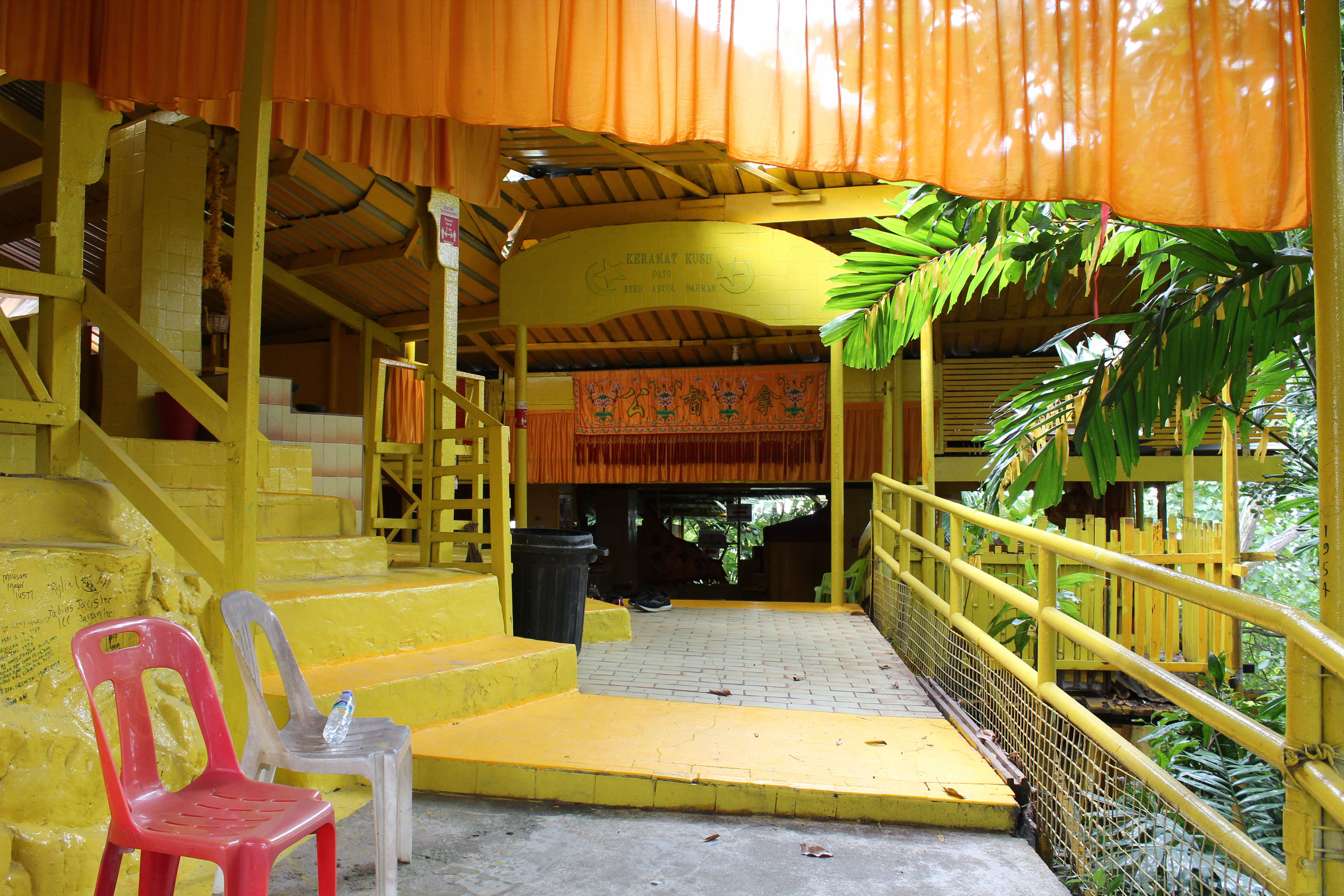 A yellow painted building with steps and a ramp, orange curtains, and tropical plants; red and white chairs in foreground.