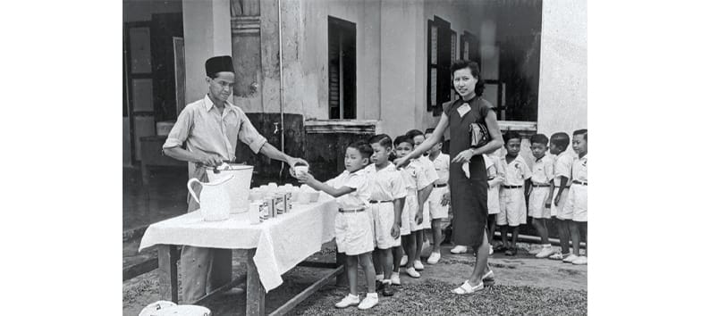 Children wearing uniforms form a line outdoors to receive drinks from an adult at a table with cups and a jug.