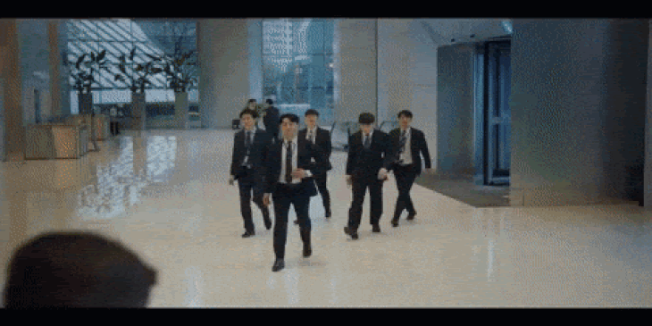 Group of men in suits walking in a modern, tiled lobby toward glass doors.