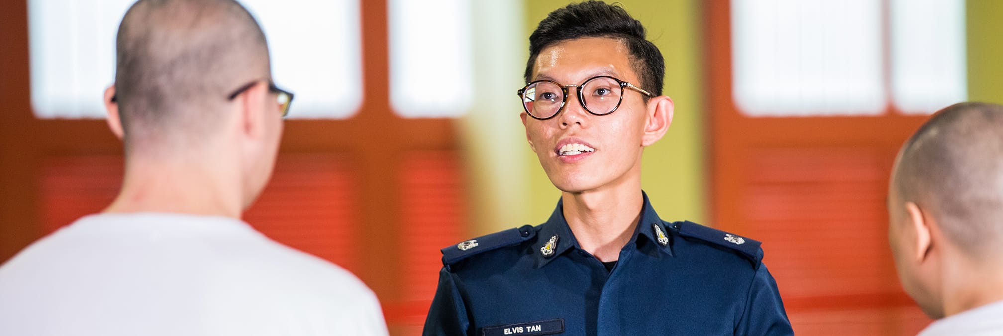 Elvis Tan in a blue uniform, speaking to two people with shaved heads.