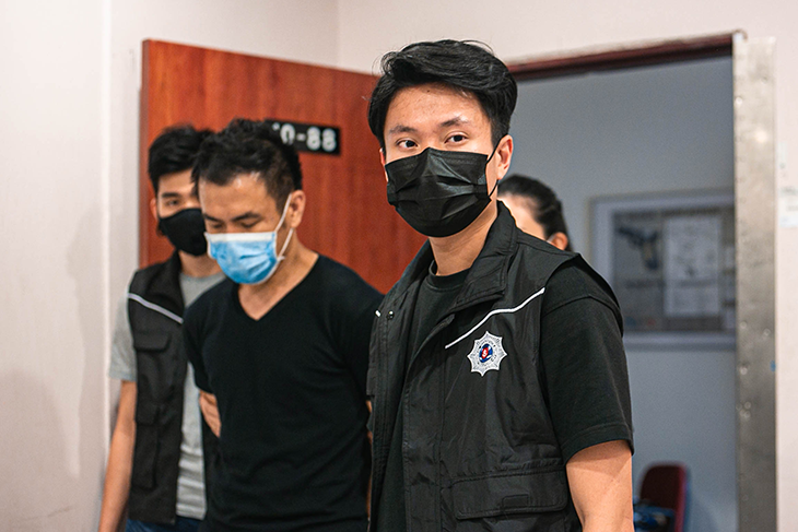 People in masks escorting a man in a black shirt, wearing vests with badges.