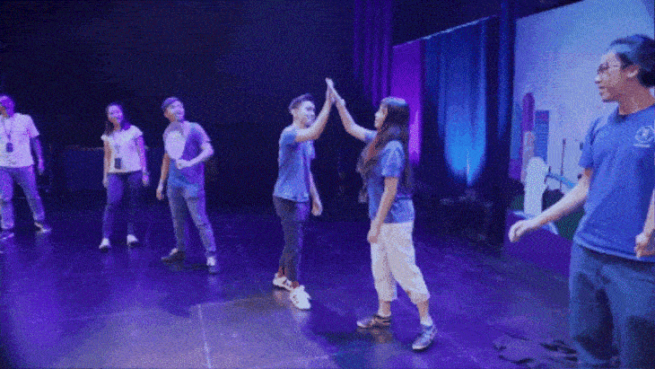 People on stage with purple lighting; two high-fiving in matching blue shirts.