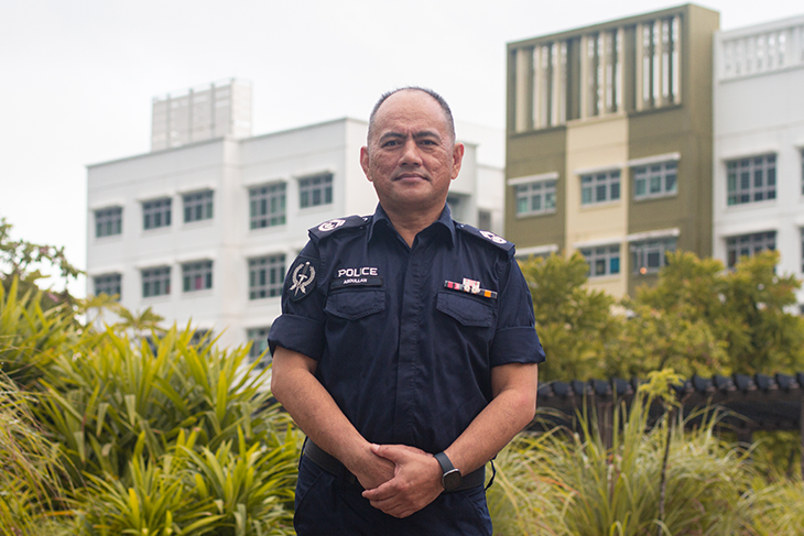 Singapore police officer in uniform stands outdoors with hands clasped. Buildings behind.