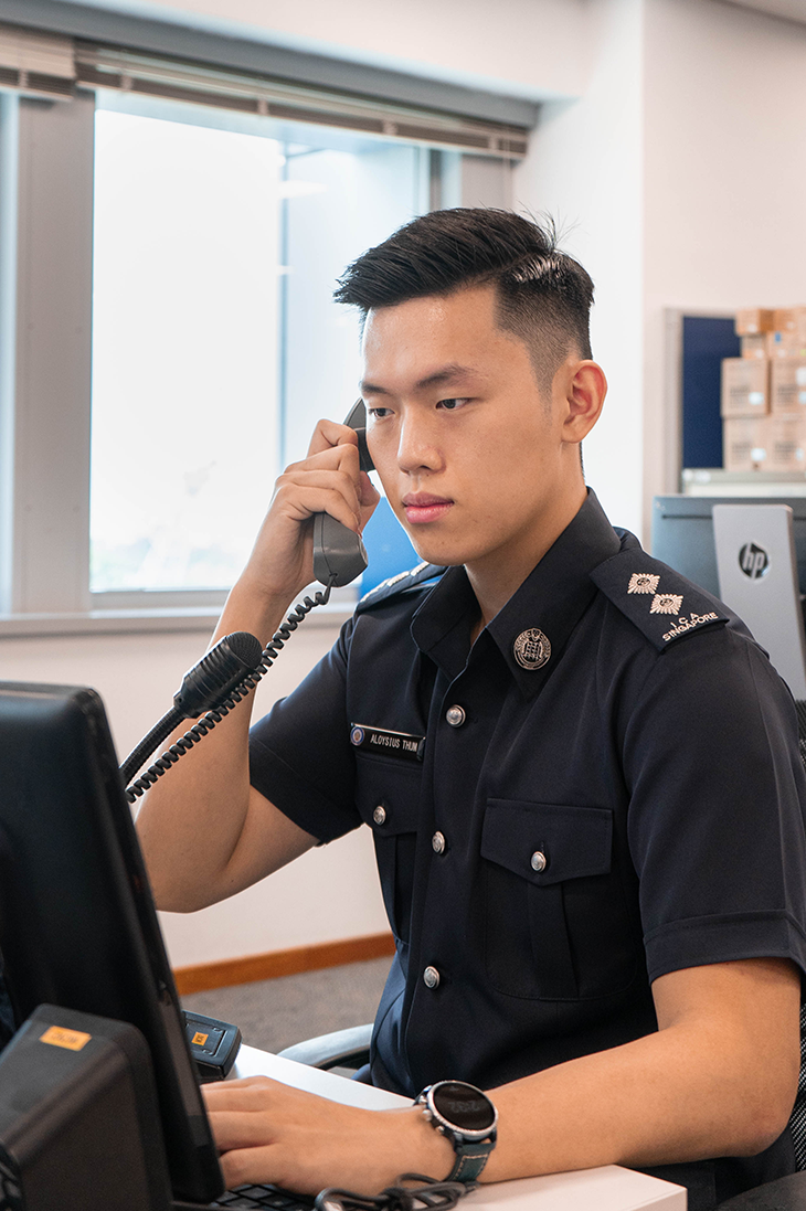 Police officer in uniform, wearing watch, on phone at computer desk near HP printer.