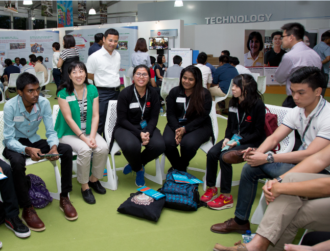 Group of people sitting in a circle on white plastic chairs in an exhibit hall. The word TECHNOLOGY is on the back wall.