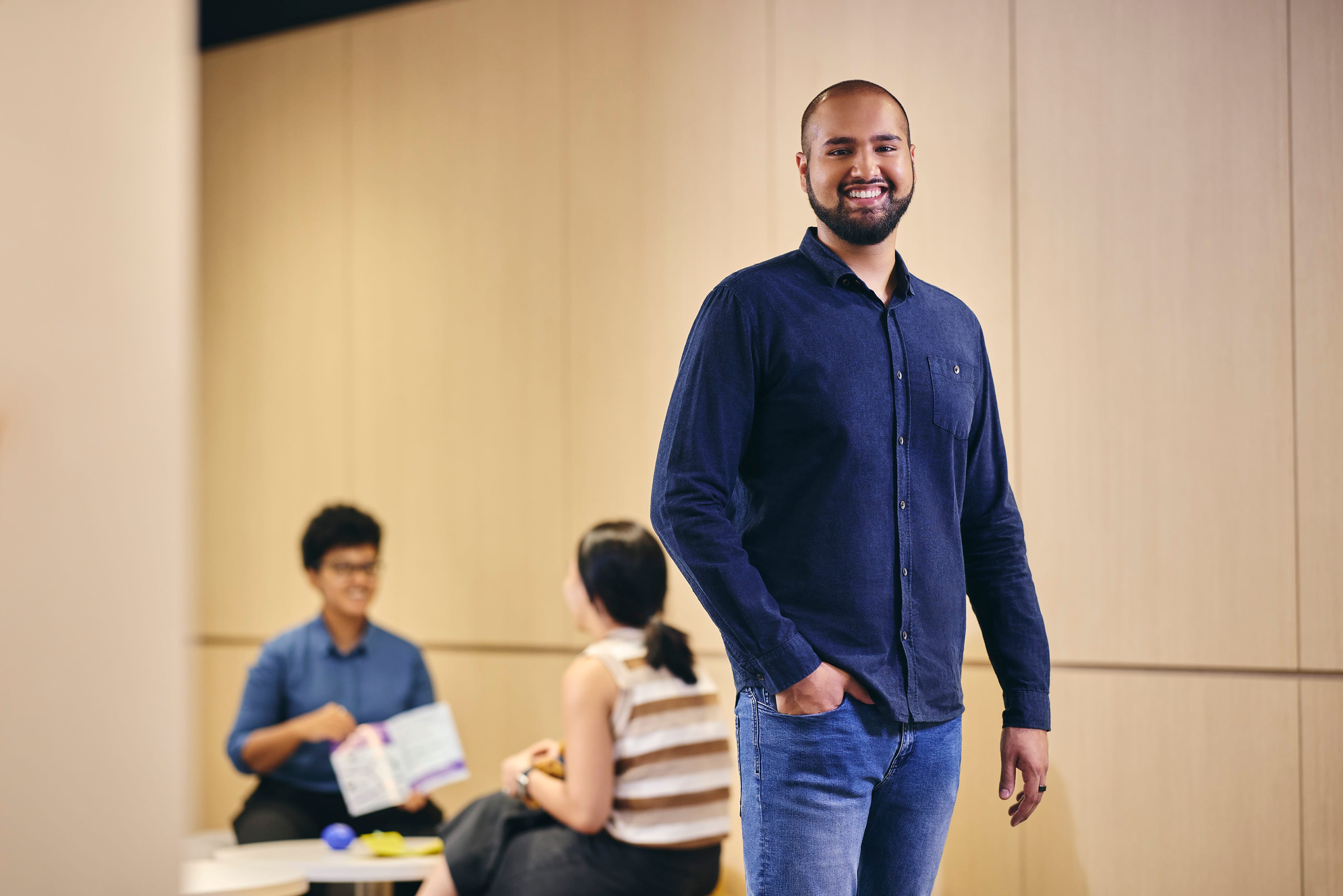 Smiling man in blue shirt and jeans, hand in pocket, with two people in background.