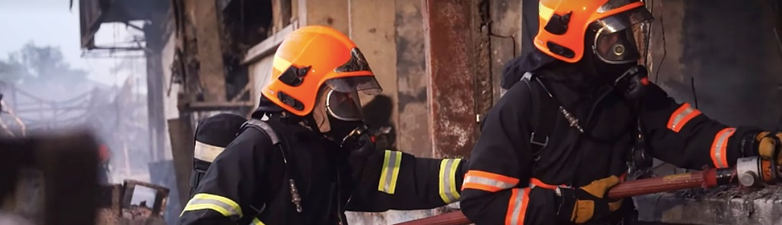 Two firefighters in full gear with orange helmets holding a hose near a damaged building.