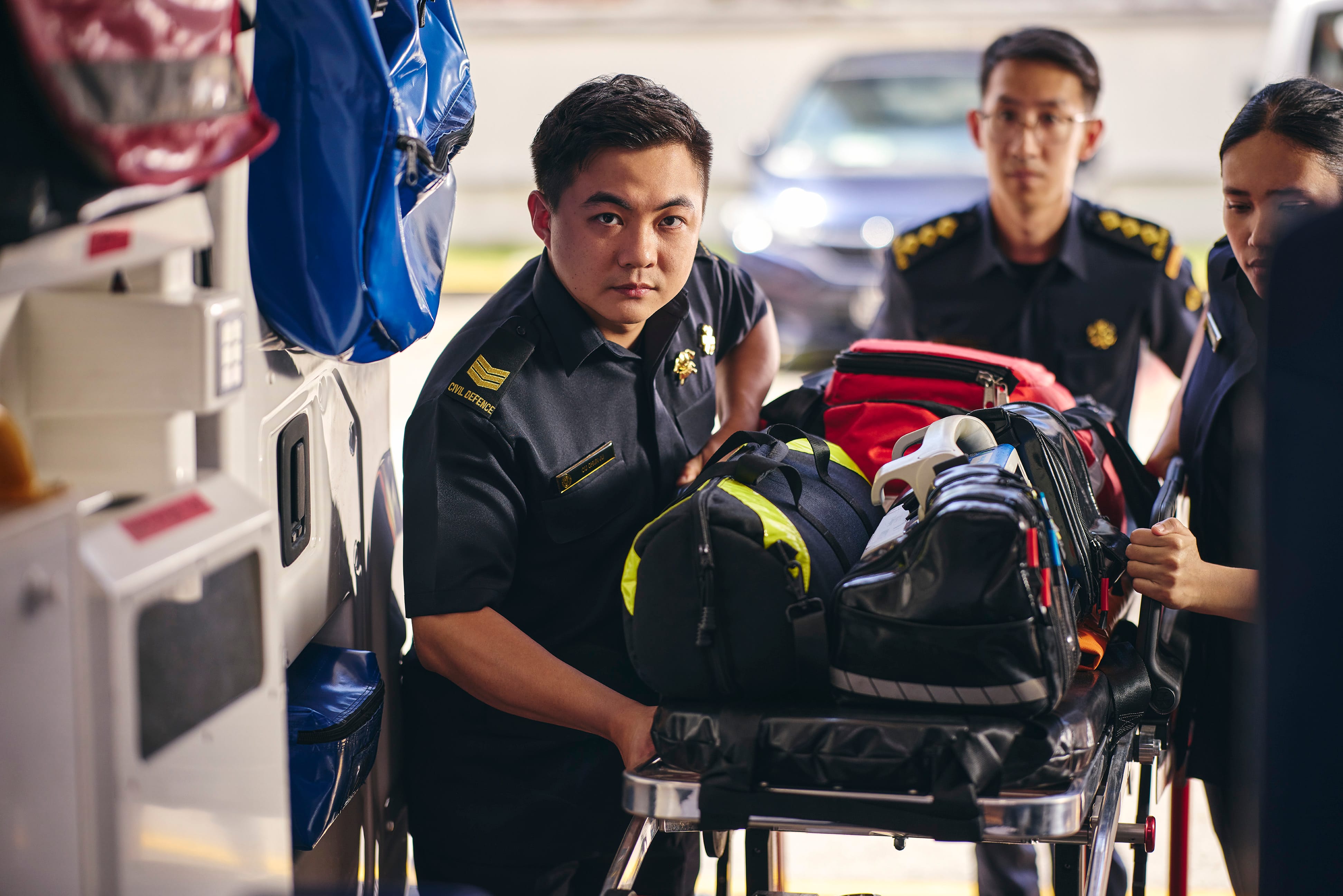 Civil Defence paramedic preparing medical equipment inside an ambulance with team members assisting.