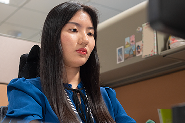 Woman with long dark hair wearing a blue blouse sitting in an office cubicle.