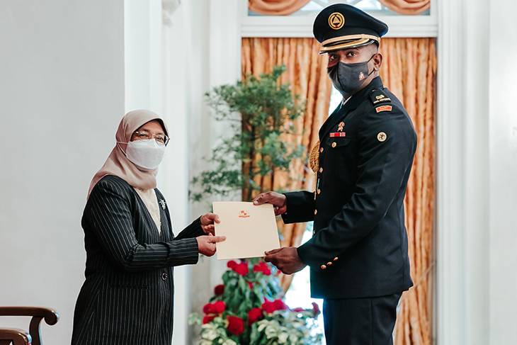 Woman in hijab presents document to officer in uniform, both wear masks.