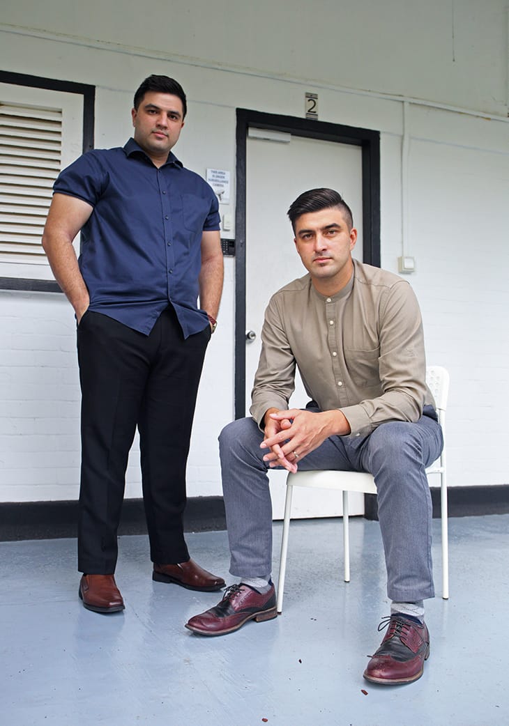 Two men pose: one stands in a blue shirt and black pants, the other sits in a tan shirt and grey pants.