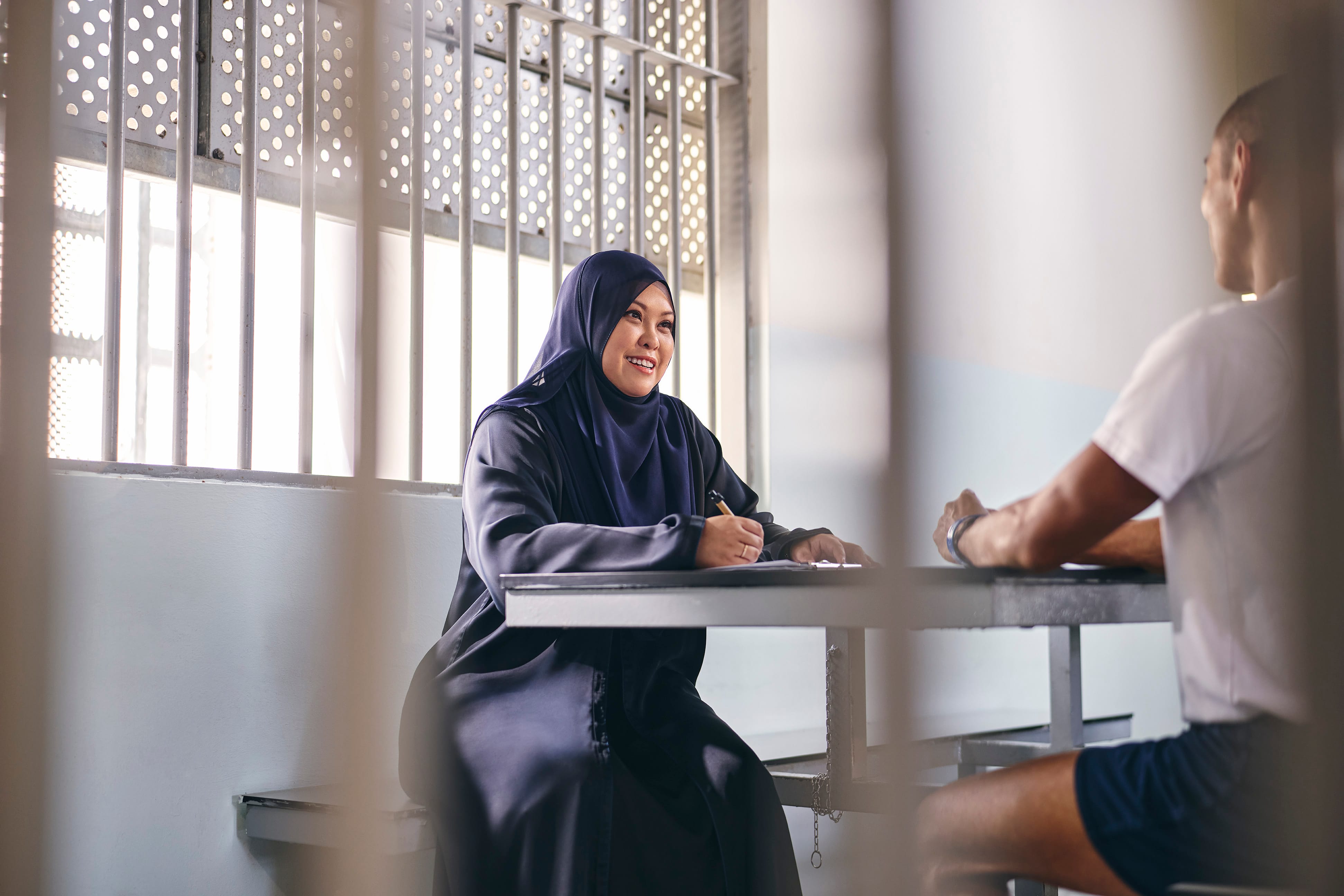 Prison officer in navy hijab smiling while speaking with an inmate across a table inside a secure facility.