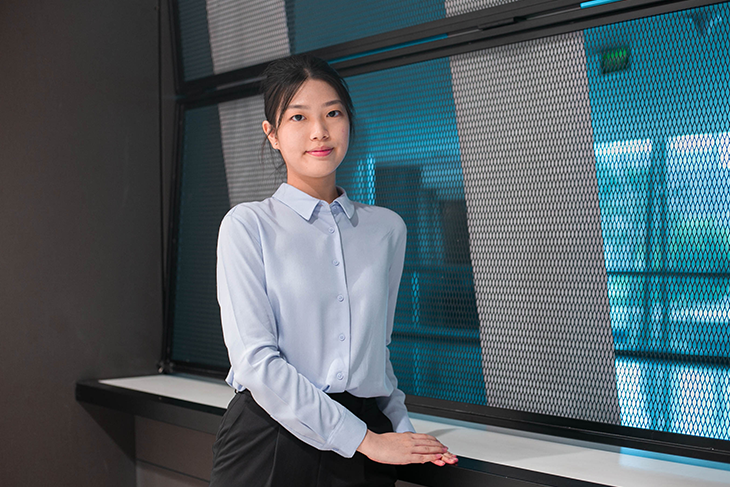 Woman in a blue shirt and black pants standing by a window with metal mesh.