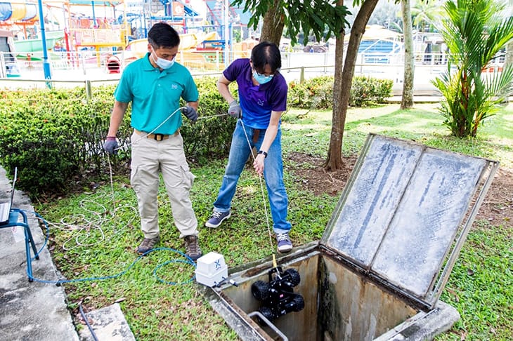 Two people using rope to lower a robot into an open concrete drain outdoors.