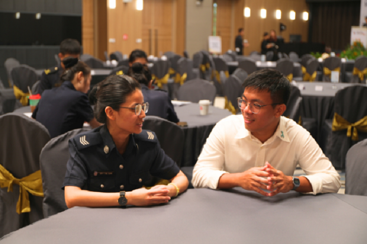 Two people seated at a table in a meeting room, one in uniform, talking.