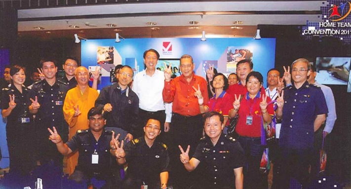 Group of people at "HOME TEAM Convention 2011" backdrop, most making peace signs.