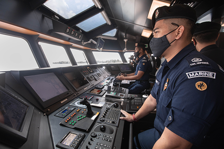 Civil Defence Marine officer in uniform and face mask operating a boat with multiple display screens.