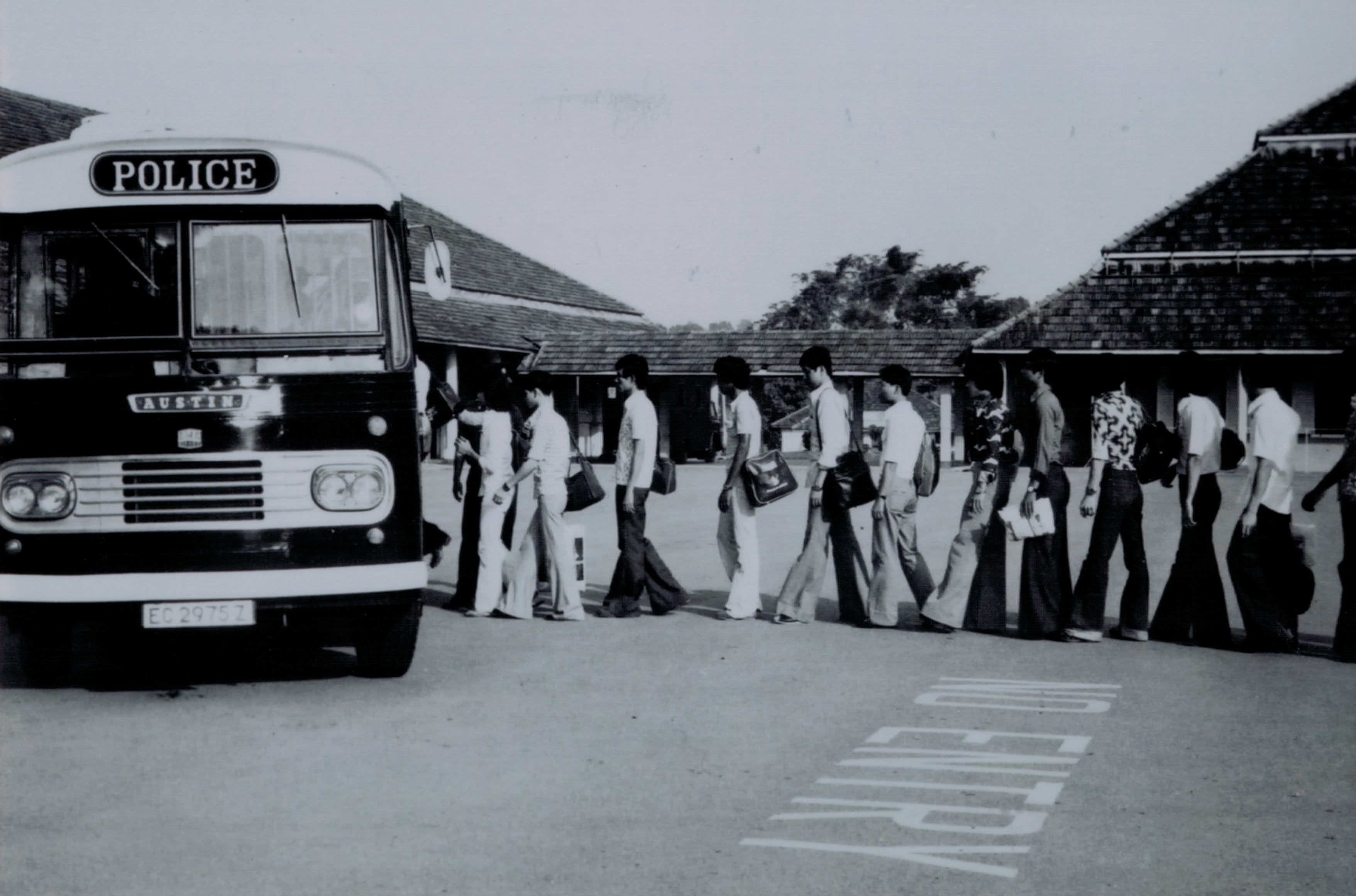 Monochrome photo of an Austin Police bus; a line of people walk toward it. "NO ENTRY" painted on the road.