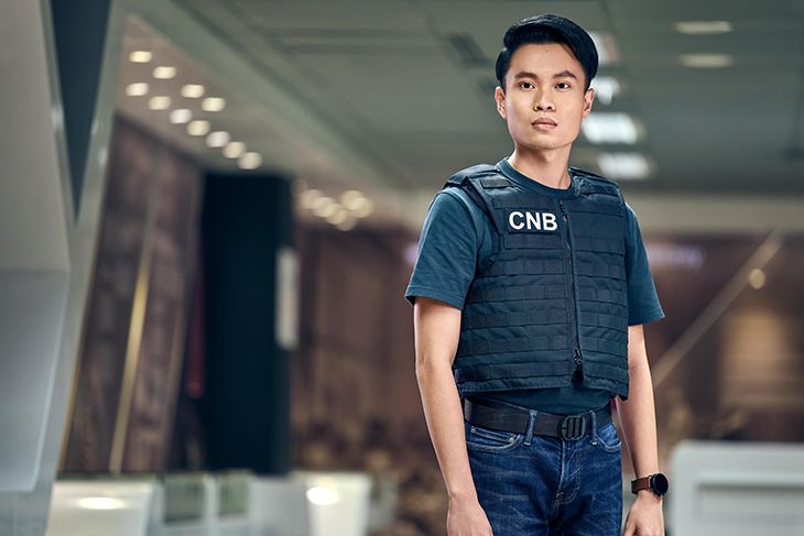 Man in CNB vest, t-shirt, jeans, and belt with smartwatch in blurred office.