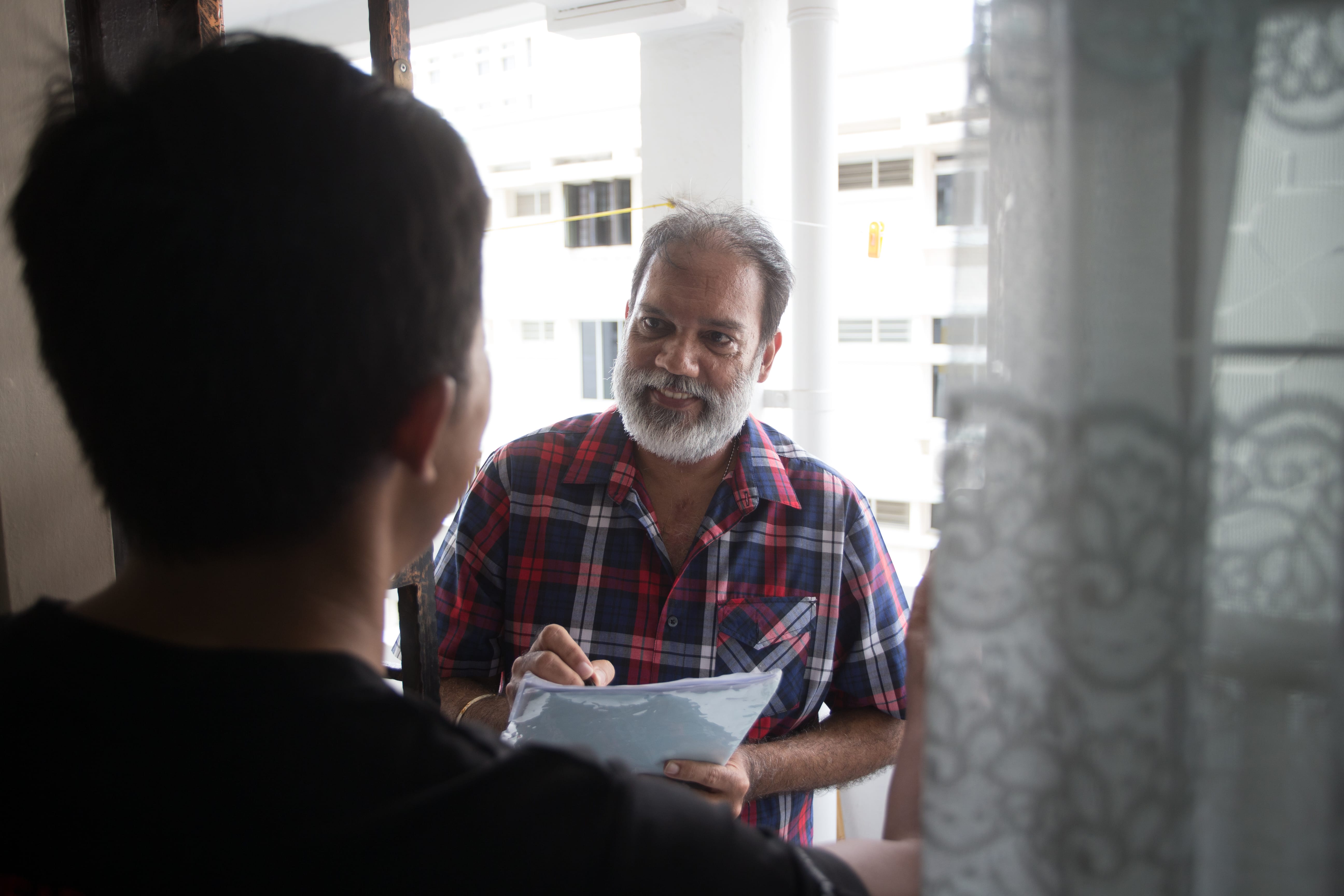 Volunteer holding a clipboard while speaking to a resident at the doorstep of an HDB flat.