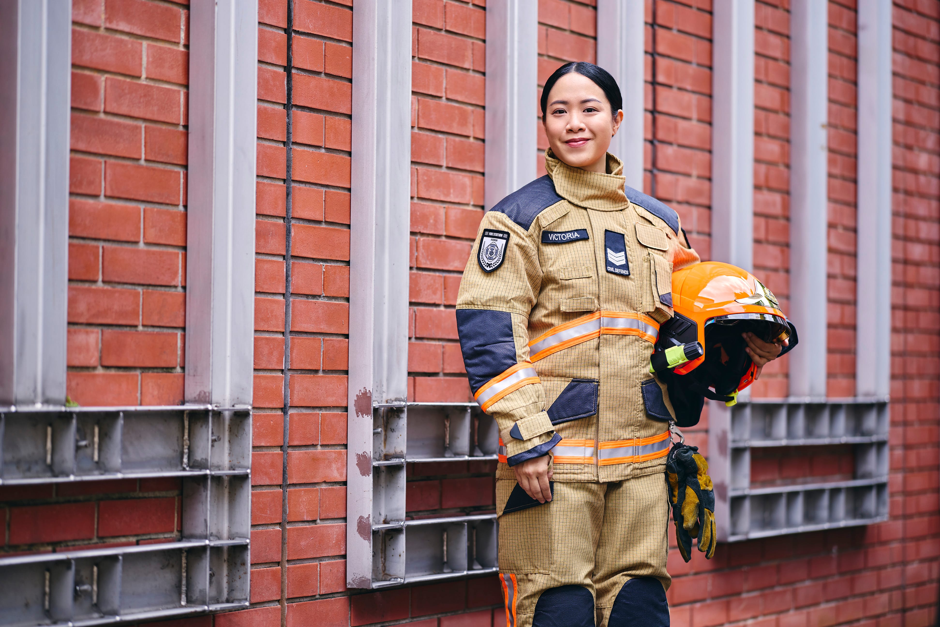 Firefighter "Victoria" in tan uniform with helmet & gloves, standing against a brick wall.