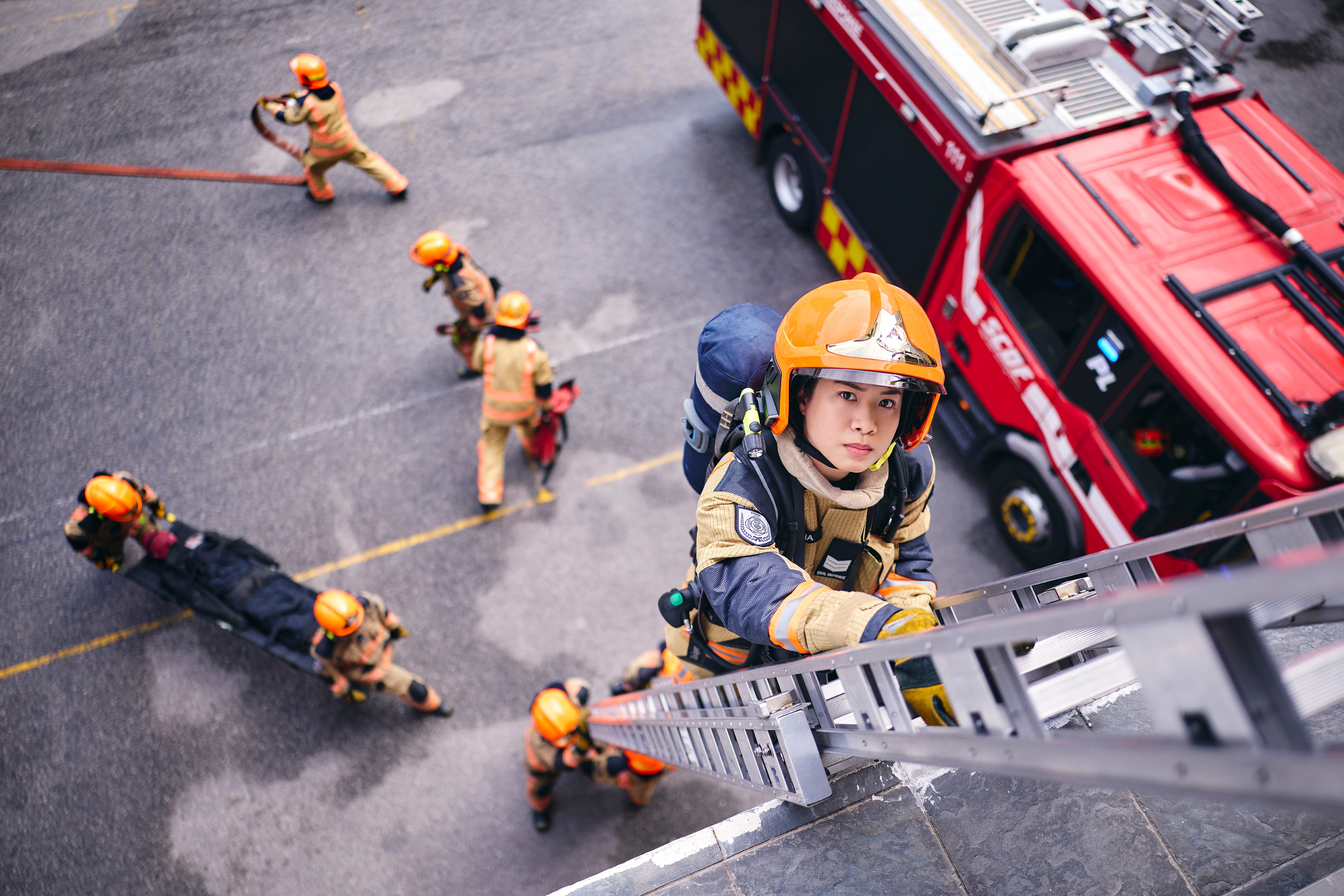 Firefighter climbing a ladder during a rescue drill, with teammates and a fire engine visible below.