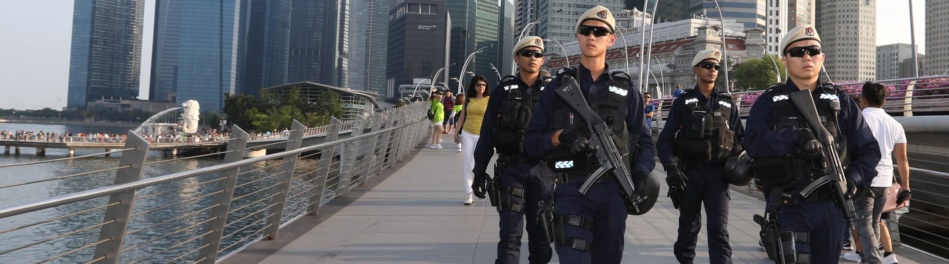 Armed officers in tactical gear walk on a bridge with city skyline and the Merlion in the background.