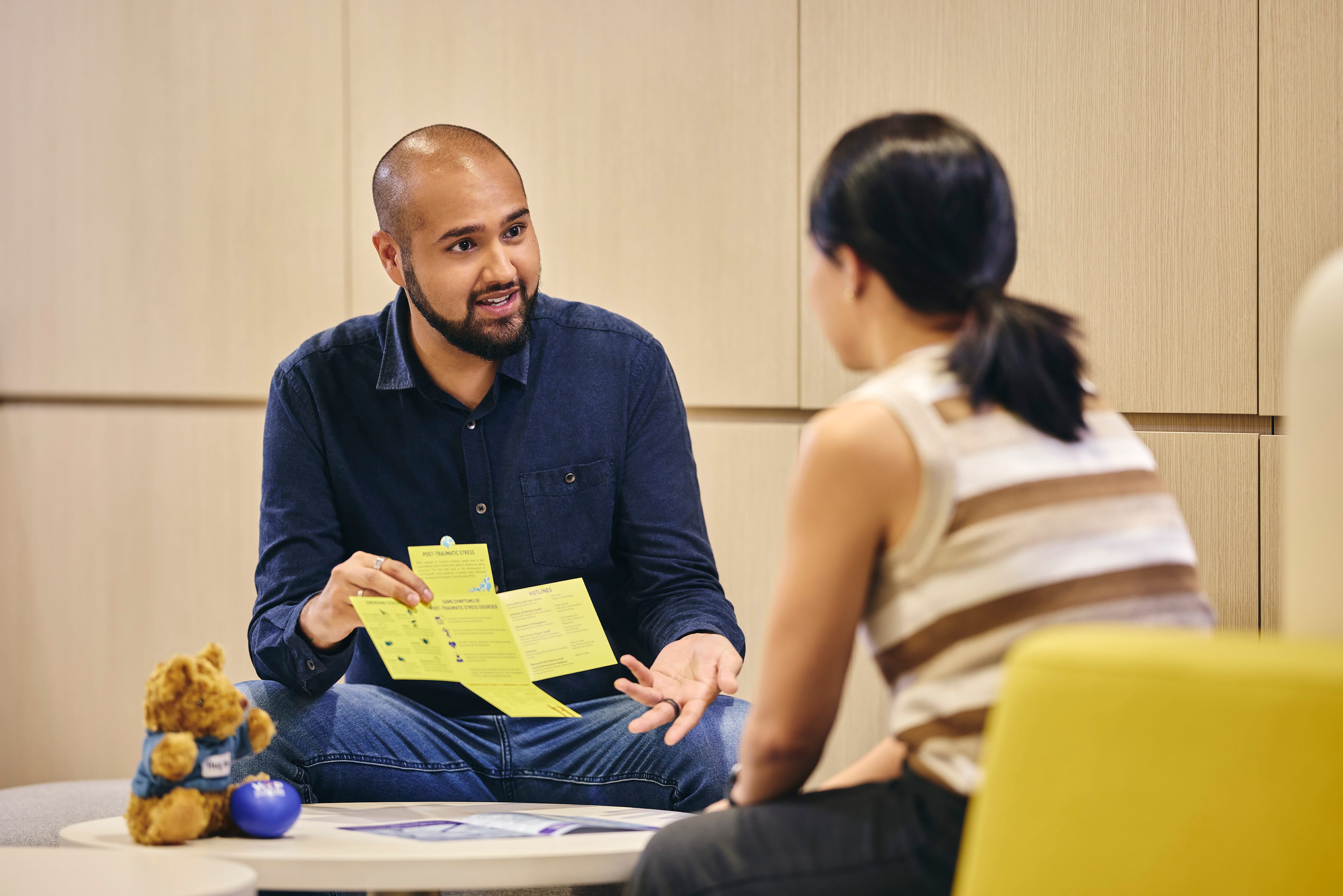 Man in a dark blue shirt sitting and explaining information from a yellow brochure to a woman in a striped top.