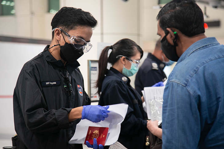 Officer in uniform examining a Malaysia passport at customs; others are waiting.