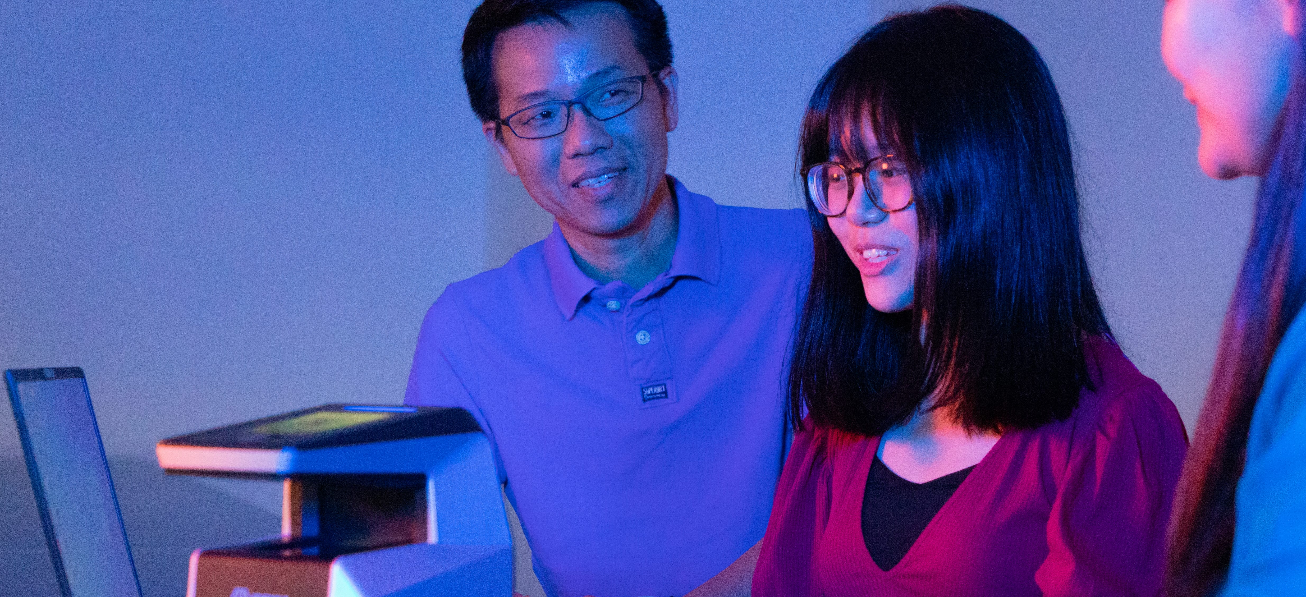 Three people gathered around a computer under blue lighting, discussing or working on a project.