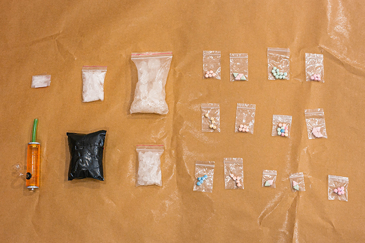 Drugs and paraphernalia on brown paper, including clear bags of crystals and pills, plus a black bag.